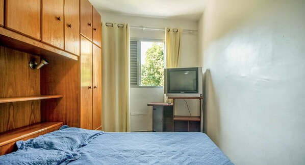 A bedroom with a wooden wardrobe, a bed with a blue blanket, a television on a wooden stand, and a window with curtains.