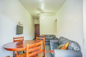 A compact living space with a dining table and chairs, a sofa with patterned cushions, a television mounted on the wall, and a ceiling fan.