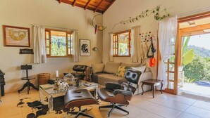 A cozy living room with a sofa, two chairs, a coffee table, and a desk. There are two windows with curtains, a hanging plant, and a view of greenery outside. - Casa em Mairinque em meio a Natureza com Piscina (Mairinque)
