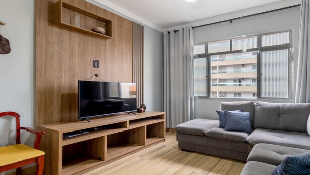 Una sala de estar con televisor de pantalla plana sobre un mueble de madera, un sofá gris, un sillón rojo y una ventana con cortinas.
