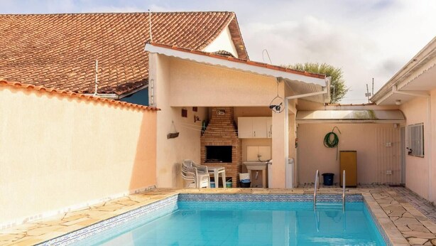 A swimming pool with a tiled deck, a brick fireplace, and a small kitchen area with a sink and cabinets. - Casa em Peruíbe com Piscina e Churrasqueira (Peruíbe)
