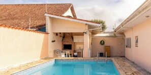 Une piscine avec une terrasse carrelée, une cheminée en briques et un petit coin cuisine équipé d’un évier et de placards.