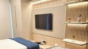 A modern hotel room with a bed, a flat-screen TV mounted on the wall, a bedside table, and a shelf with decorative items.