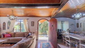 Un salón espacioso con techo de madera, un sofá grande, una zona de comedor con mesa y sillas de madera, y vistas a un jardín a través de un arco.