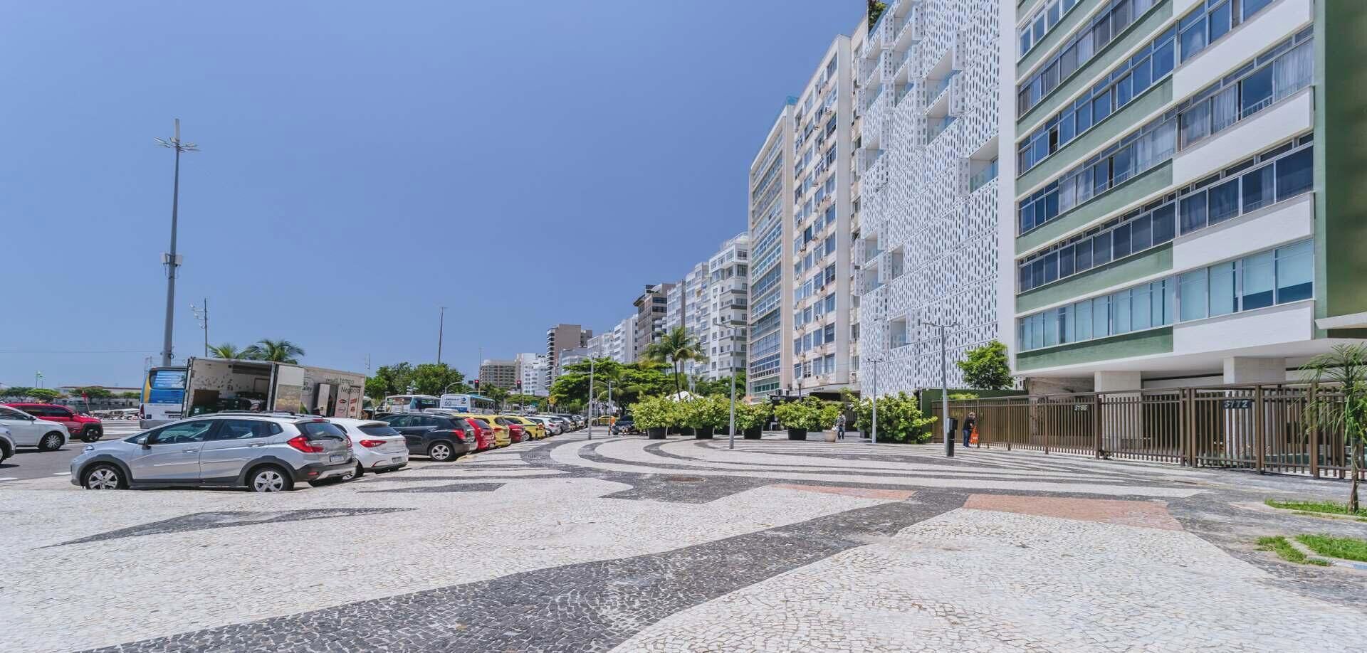A modern urban street with a parking lot, lined with multi-story buildings featuring balconies and a mix of glass and metal facades.
