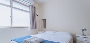 A bedroom with a bed, a nightstand, a lamp, a window with curtains, and a mirror on the wall.