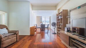 A living room with a brown sofa, a dining table with chairs, a TV stand with a flat-screen TV, and a standing fan.