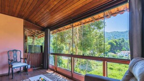 Una habitación con ventanas amplias, techo de madera, televisor, sofá y una silla.