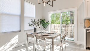 Un coin repas avec une table dressée pour quatre personnes, entourée de chaises blanches, un vase avec des plantes vertes et un luminaire suspendu moderne.