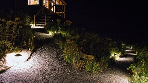 A house with a wooden staircase leading to a second floor, surrounded by a gravel path and illuminated by ground lights.