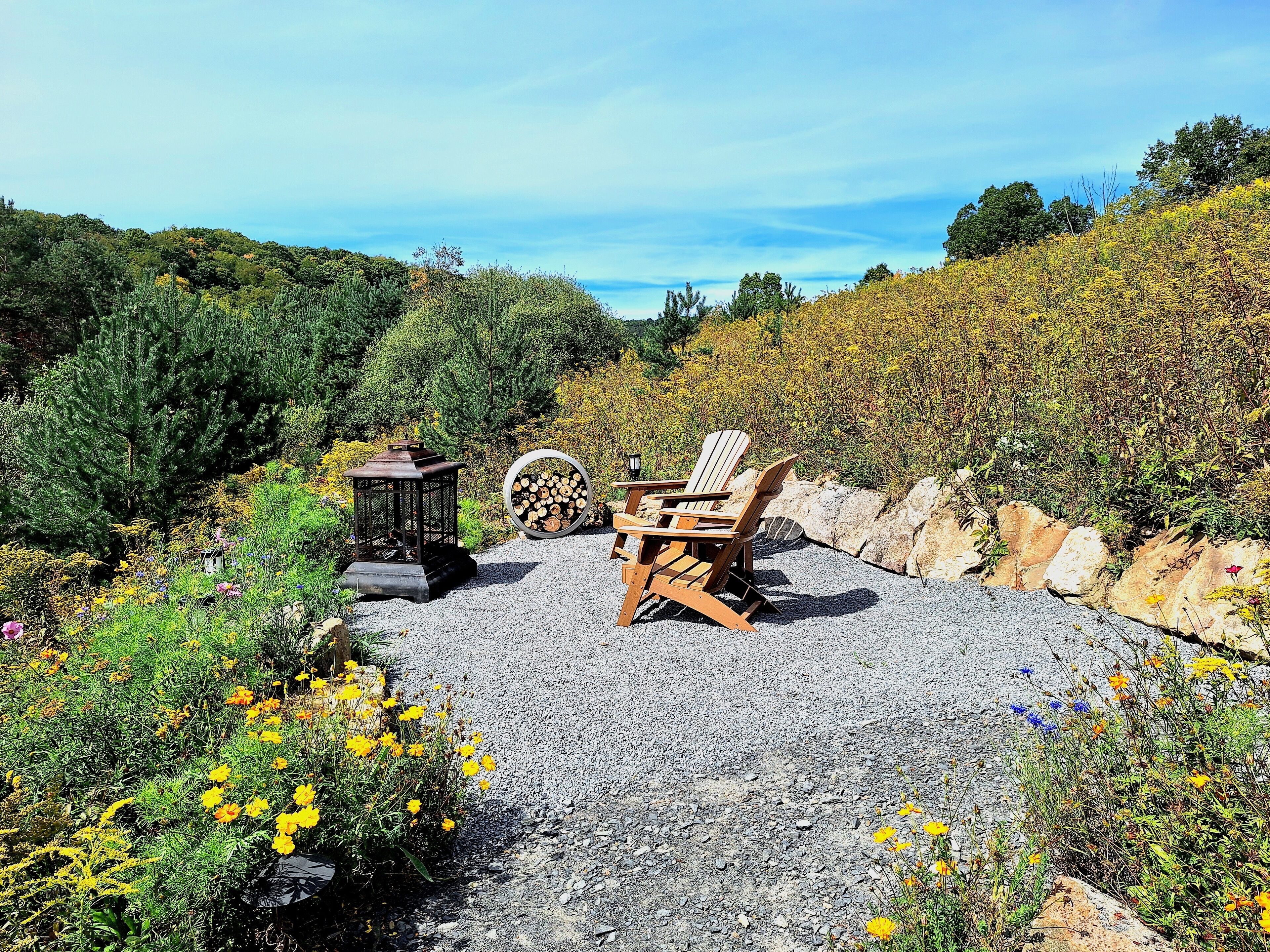 A gravel patio with Adirondack chairs, a lantern, and a stack of firewood.