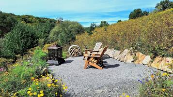 A gravel patio with Adirondack chairs, a lantern, and a stack of firewood.