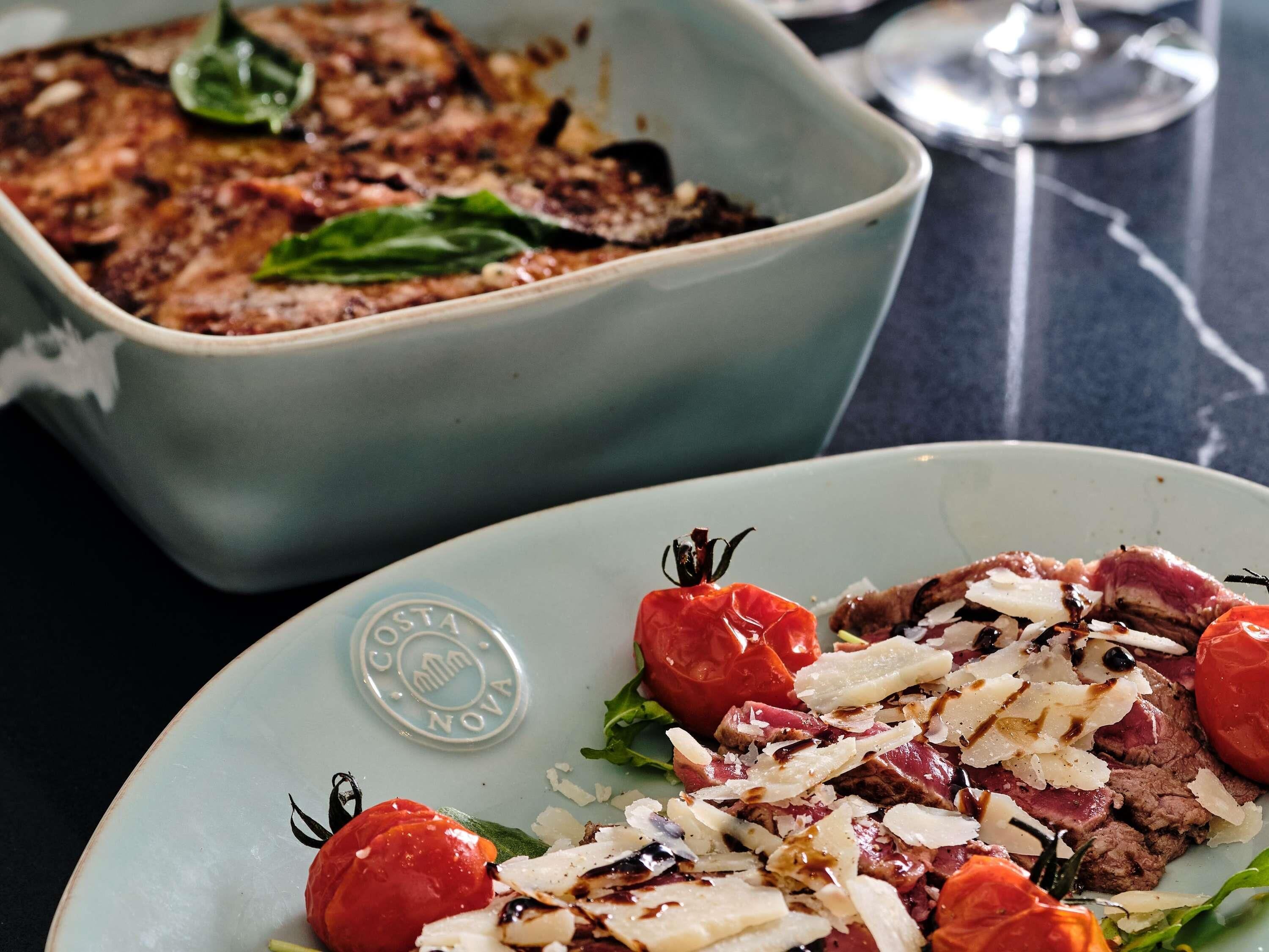 Une assiette de nourriture avec des tomates et du fromage, un plat en arrière-plan et un verre de vin sur une table.