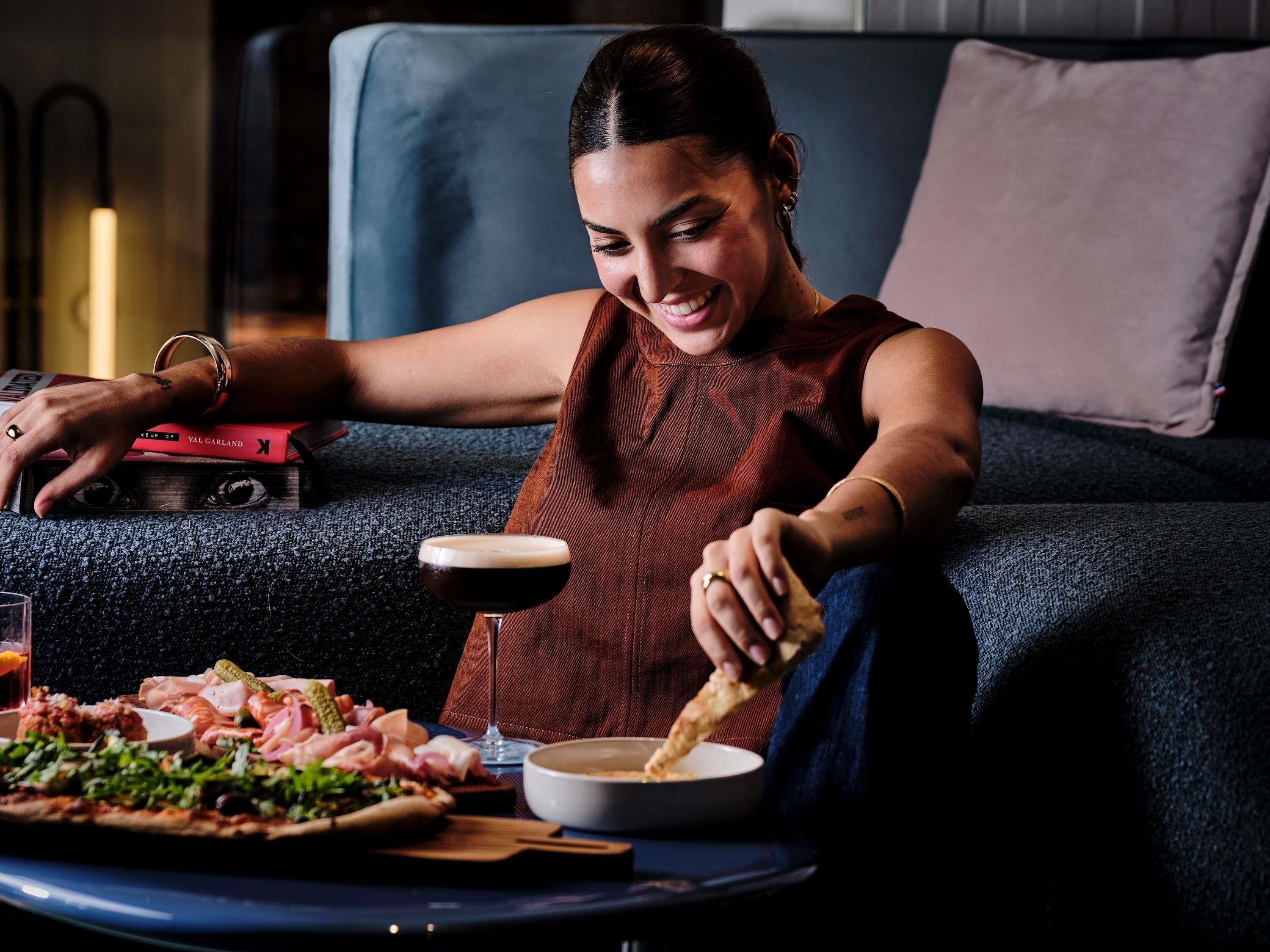 A person sitting on a couch with a plate of food, a drink, and a book titled 'VAL GARLAND' on the couch.