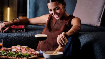 A person sitting on a couch with a plate of food, a drink, and a book titled 'VAL GARLAND' on the couch.
