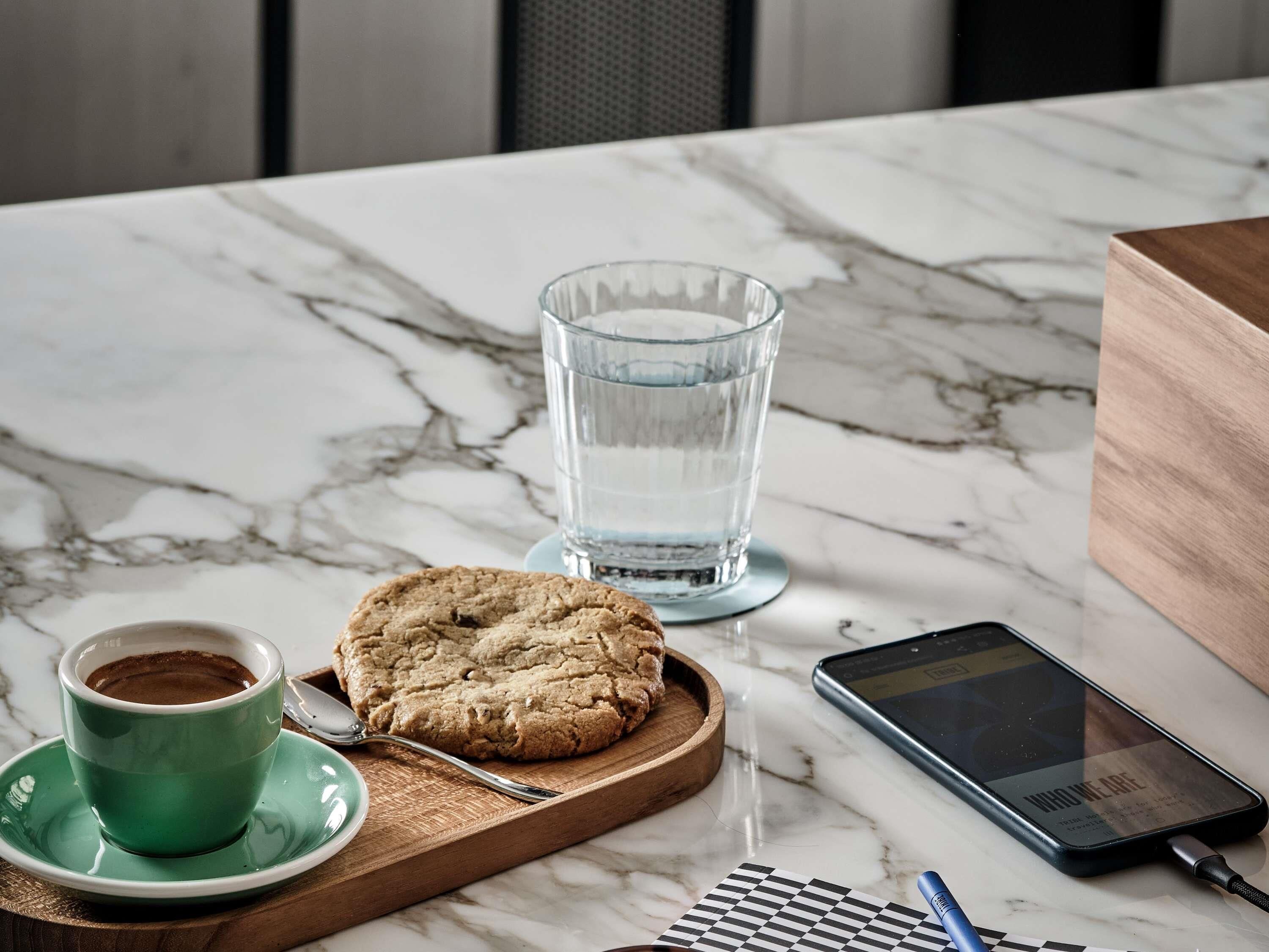 Una mesa de mármol con un vaso de agua, una galleta, una taza de café, un teléfono inteligente y un cuaderno.
