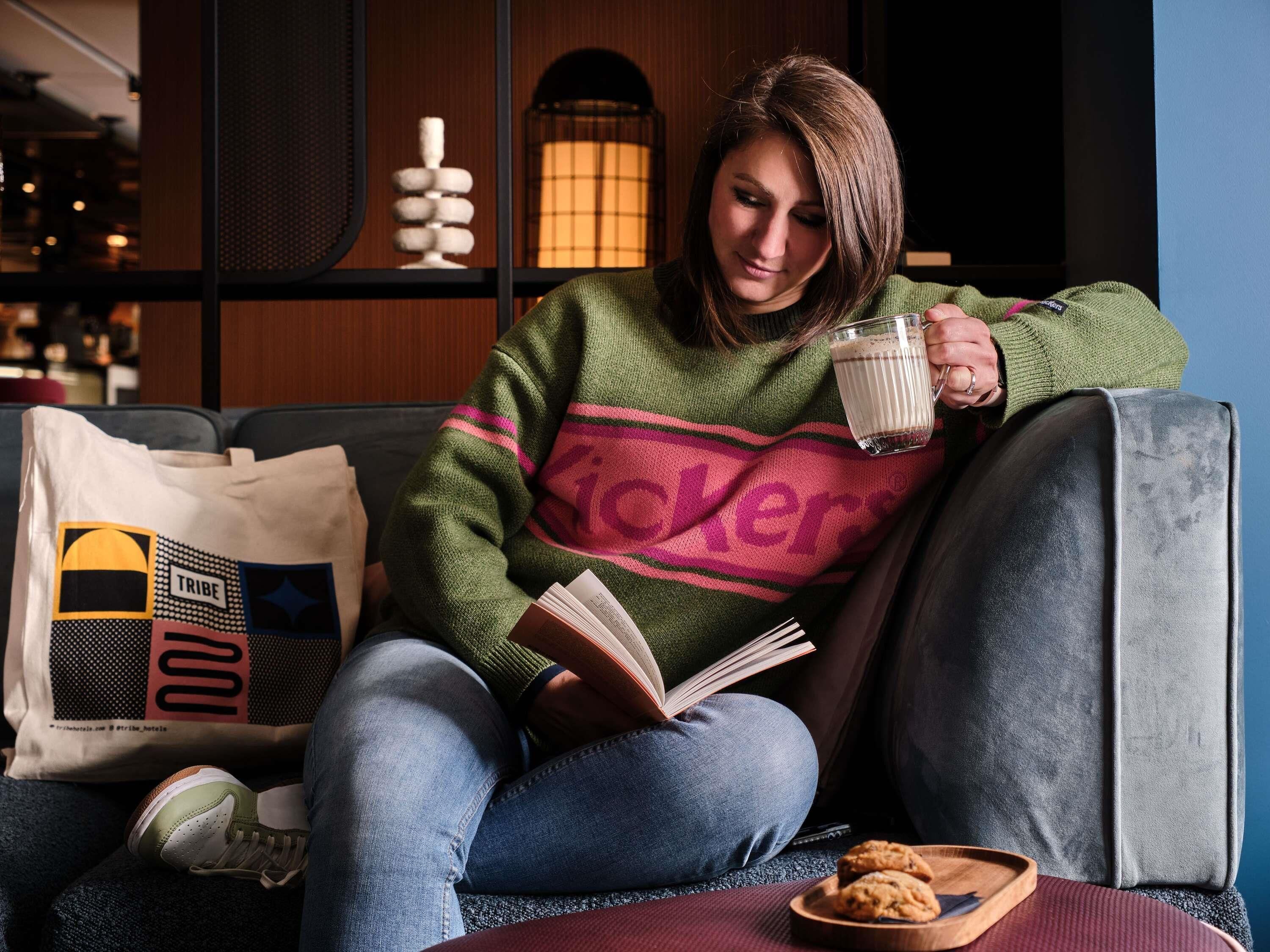 A person sitting on a sofa with a book, a glass of milk, and a cookie tray.