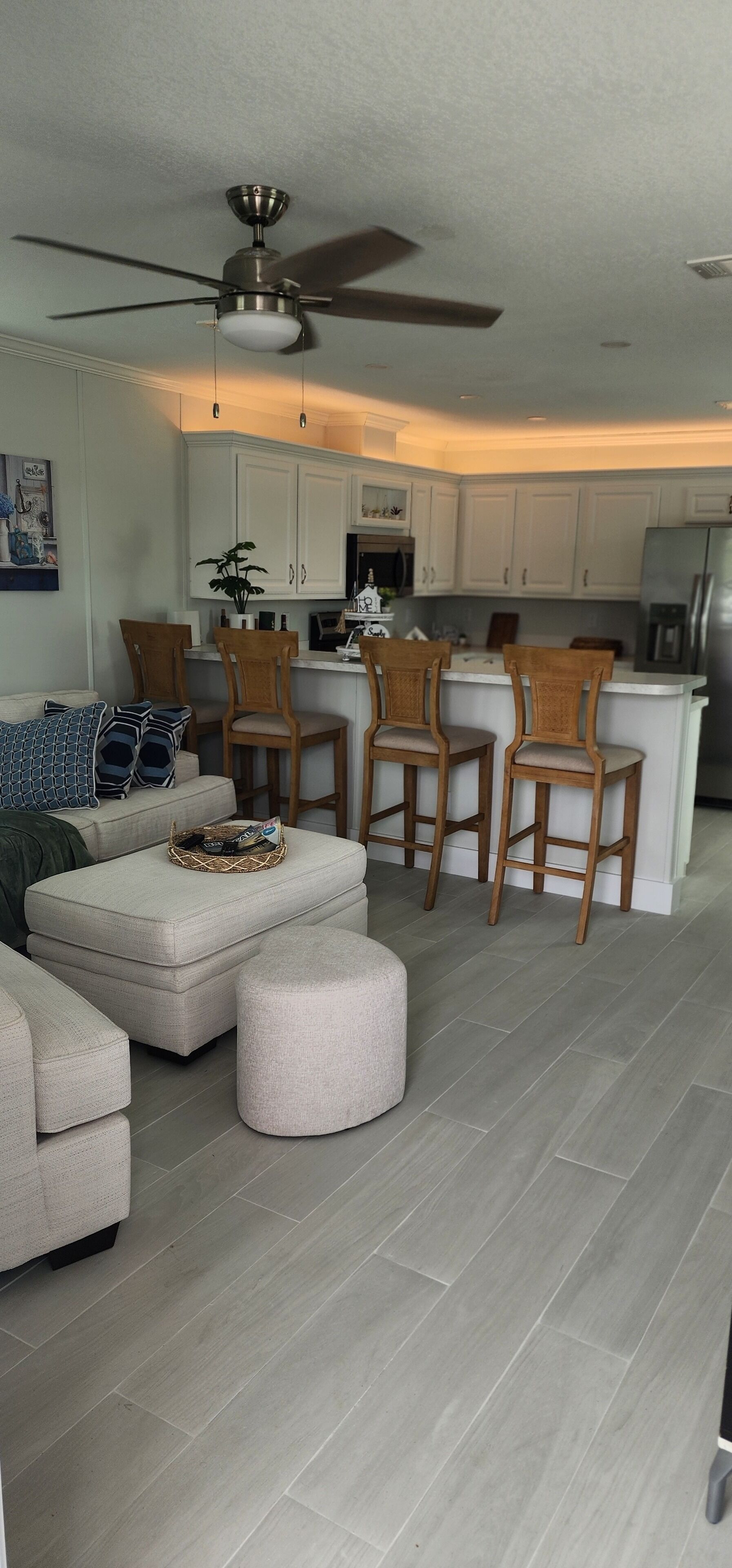 A modern living room with a sofa, ottomans, and wooden chairs. A kitchen with white cabinets and a bar area is visible in the background.