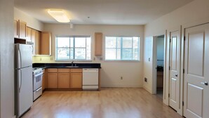 A compact kitchen with wooden cabinets, a built-in oven, a refrigerator, and a dishwasher. There are two windows providing natural light, and a door leading to another room. - Live Close to Everything! (Oakland)