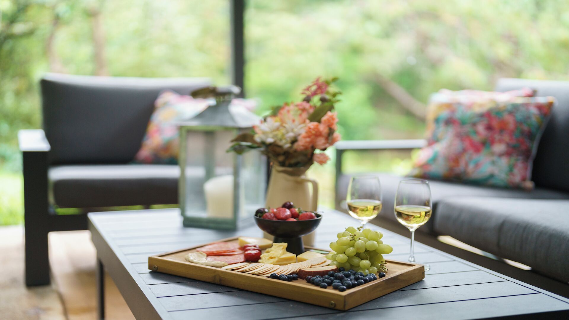 A wooden table with a charcuterie board, a bowl of grapes, and two glasses of white wine.