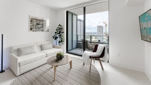 A modern living room with a white sofa, a round coffee table, a potted plant, and a view of a cityscape through large windows. - 601 RESIDENCES WONDERFUL 1BR UNIT #3203 (Miami)
