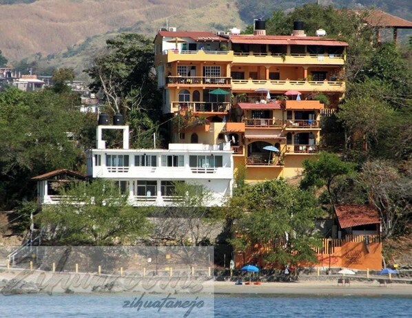 Um edifício de vários andares com varandas, uma estrutura branca em frente e uma praia ao fundo.