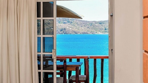 Habitación con puerta de vidrio corredera que da a un balcón con mesa y sillas, con vistas a una masa de agua y colinas.