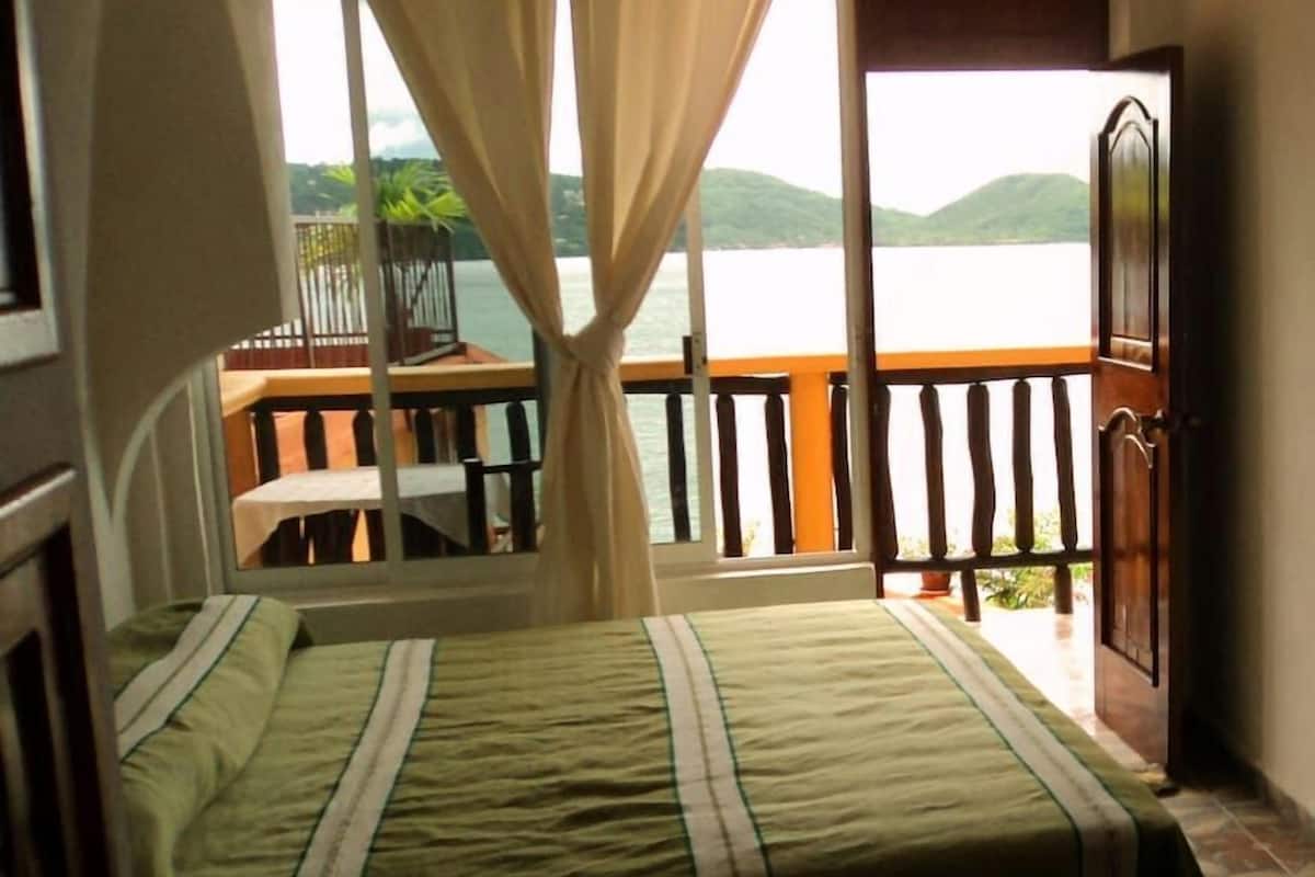 A bedroom with a bed, a balcony with a view of the water, and a door.