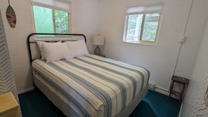 Une chambre à coucher avec un lit, deux fenêtres, une lampe de chevet et un petit tabouret en bois.