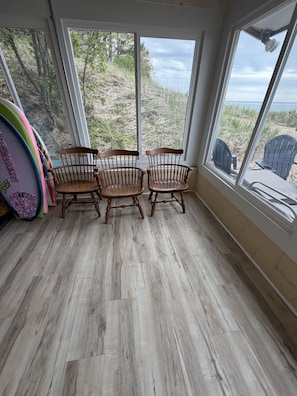 Quarto com piso de madeira, duas cadeiras de madeira, uma mesa e uma prancha de surf.