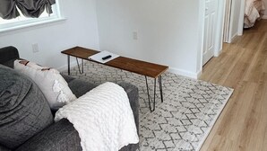 Una sala de estar moderna con un sofá gris, una manta blanca, una mesa de centro de madera y un tapete estampado. Un televisor de pantalla plana está montado en la pared, y se ve una ventana con cortinas.