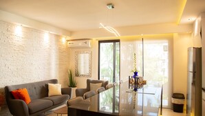 Una sala de estar moderna con un sofá, sillas, una mesa de comedor de cristal y una zona de cocina con un refrigerador.