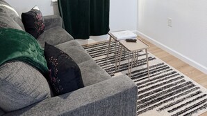 Un salon avec un canapé gris, des coussins verts et noirs, un téléviseur noir fixé au mur et un tapis à motifs sur le sol.
