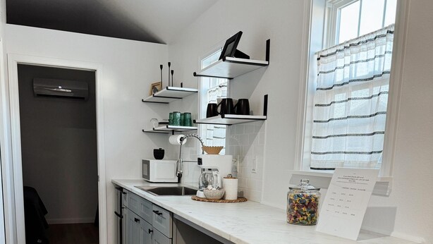 Une cuisine moderne avec un plan de travail blanc, des meubles gris et une fenêtre ornée de rideaux à rayures. On y trouve trois tabourets blancs et une machine à café posés sur le plan de travail.
