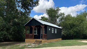 Une petite cabane à un seul étage, dotée d’un porche et entourée d’arbres, avec un chemin de gravier.