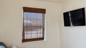 A living room with a sofa, a coffee table, a TV mounted on the wall, and a window with blinds.