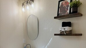 A bathroom with a white sink, mirror, and toilet. There are shelves with a framed picture and a plant.