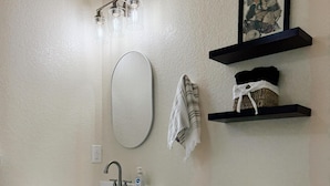 Een badkamer met een wit wastafel, spiegel en toilet. Boven het toilet bevinden zich twee planken, waarvan er een een fotolijst heeft en de andere handdoeken.