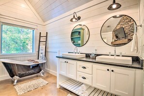 Une salle de bain avec une baignoire sur pieds, deux lavabos, des meubles blancs, un miroir et une fenêtre avec des stores.