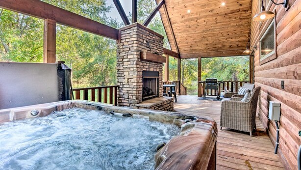 A hot tub, stone fireplace, wicker furniture, and a wooden deck with a view of trees. - Romantic King Retreat:Hot Tub-Firepit-Deck-Dogs-Sleeps4 (Broken Bow)