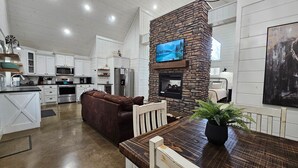 A spacious kitchen with white cabinetry, a stone fireplace, a large flat-screen TV, and a dining area with a wooden table and brown leather couch. - Romantic King Retreat:Hot Tub-Firepit-Deck-Dogs-Sleeps4 (Broken Bow)