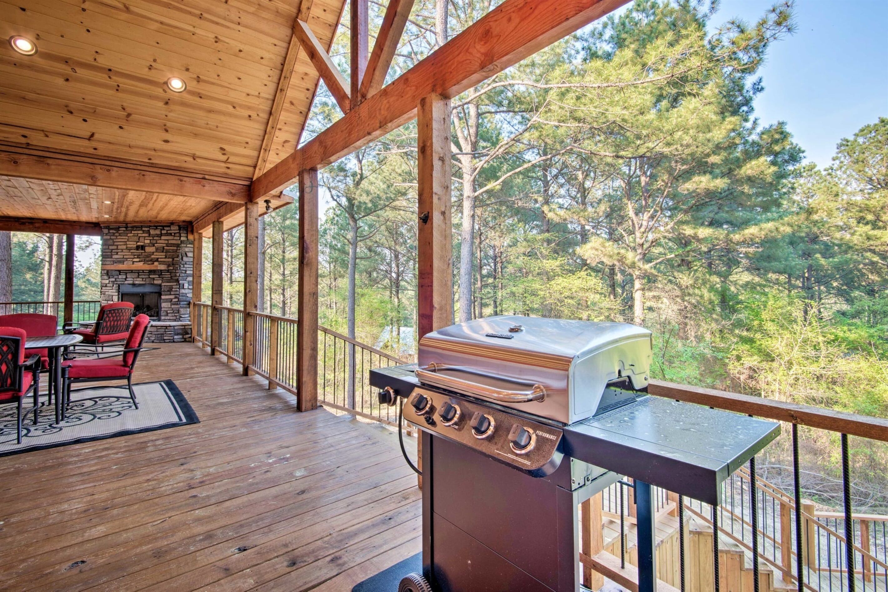 A covered outdoor deck with a stone fireplace, a grill, and a dining area with red chairs.