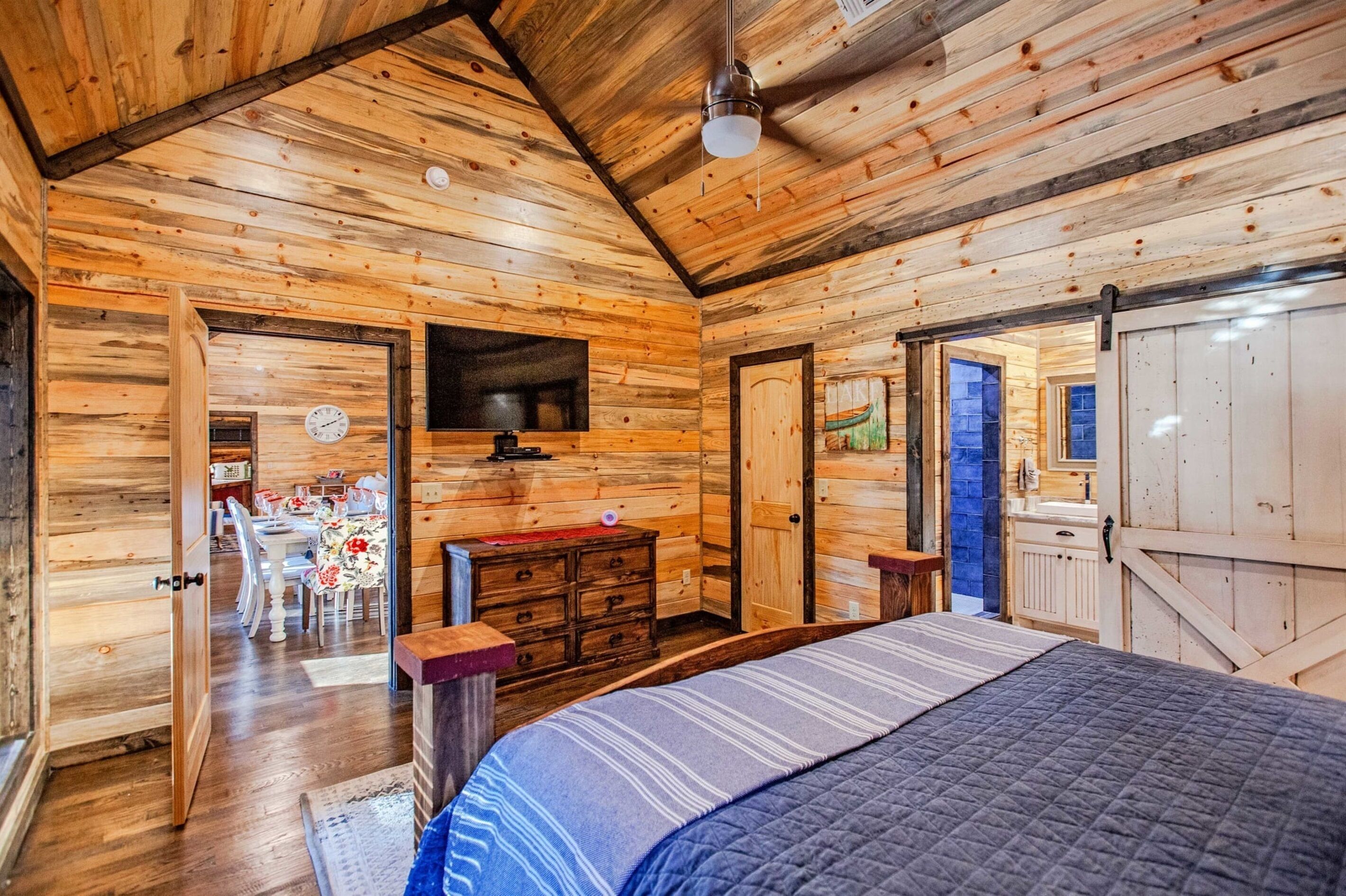 Een slaapkamer in een blokhut, met een bed, een dressoir en een eethoek die door een open deur zichtbaar is.