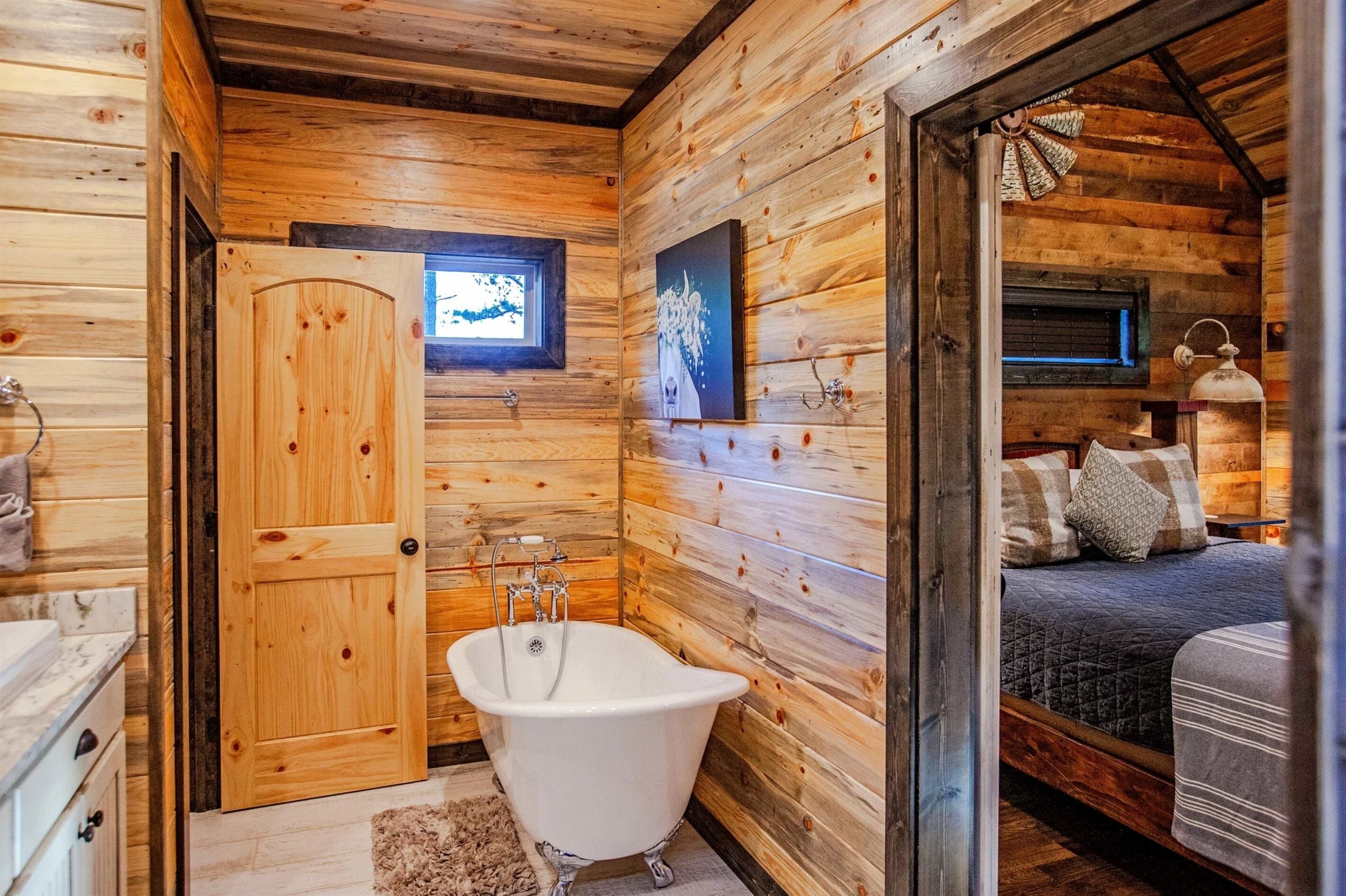 A wooden cabin interior with a wooden door, a white bathtub, a bed with pillows, and a small window.