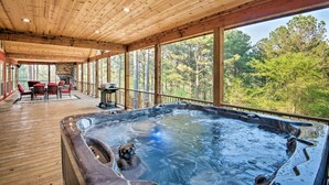 Una terraza de madera con un jacuzzi, mobiliario exterior y ventanas amplias que dan a un bosque.