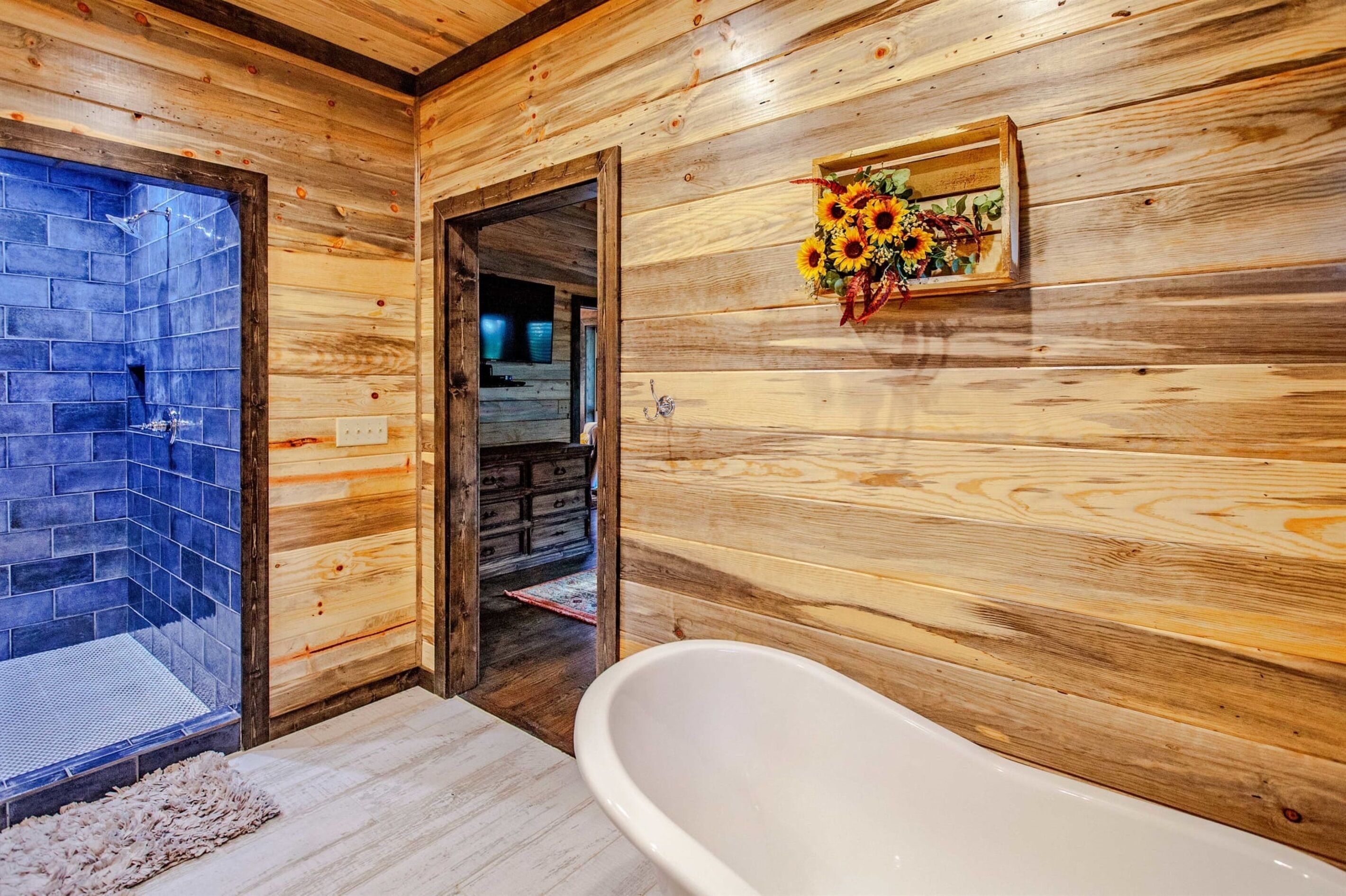 Interieur van een houten hutje, met een douche met stenen tegels, een witte badkuip en een houten deur die naar een andere kamer leidt.