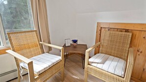 Zwei Korbstühle mit weißen Kissen, ein kleiner Holztisch und ein Fenster mit Vorhängen.