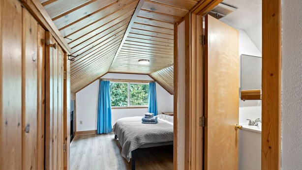 Ein Schlafzimmer mit schrägem Dach, einem Bett, einem Fenster mit Vorhängen und einer Holztür.