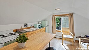 Eine kleine Küche mit Herd, Spüle und modernen Geräten. Es gibt einen Essbereich mit einem Stuhl und einem Tisch sowie ein Fenster mit Vorhängen.