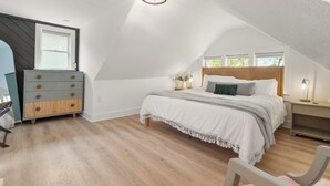 Ein Schlafzimmer mit schrägverdeckter Decke, einem Bett mit Holz Kopfteil, einer Kommode und einem Nachttisch mit Lampe.